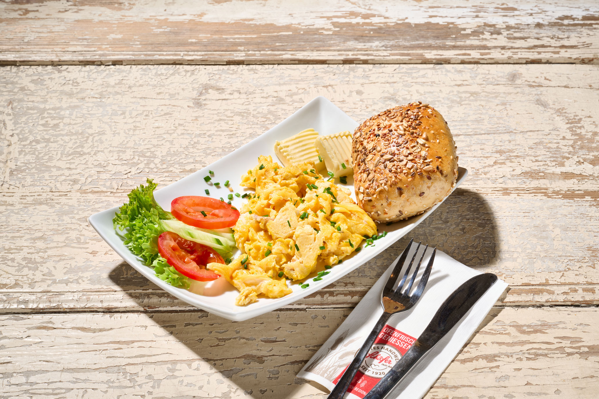 Rustikaler Teller mit Rührei, Butter, Salat und einem Körnerbrötchen auf hellem Holz