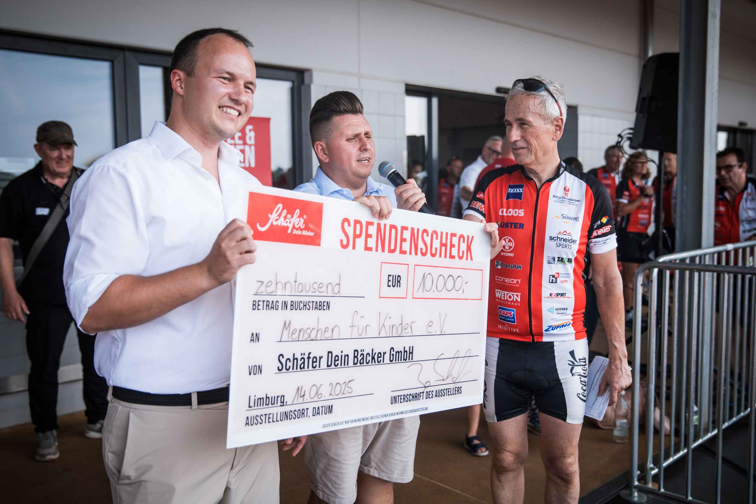 Spendenscheck-Übergabe an „Menschen für Kinder e.V.“ Drei lächelnde Personen stehen auf einer Bühne, zwei von ihnen halten gemeinsam einen überdimensionalen Spendenscheck über 10.000 € mit dem Logo „Schäfer Dein Bäcker“. Einer der Männer spricht in ein Mikrofon, im Hintergrund sind Zuschauer und Radfahrer zu sehen.