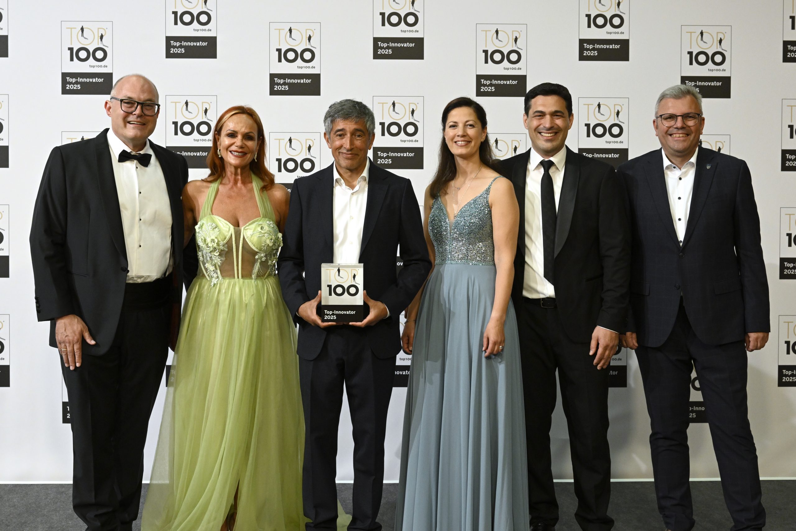 TOP 100-Innovator 2025 Auszeichnung Sechs Personen in festlicher Abendgarderobe vor einer Fotowand mit „TOP 100 Top-Innovator 2025“-Logos, der in der Bildmitte stehende Mann hält das „TOP 100“-Trophäenschild.