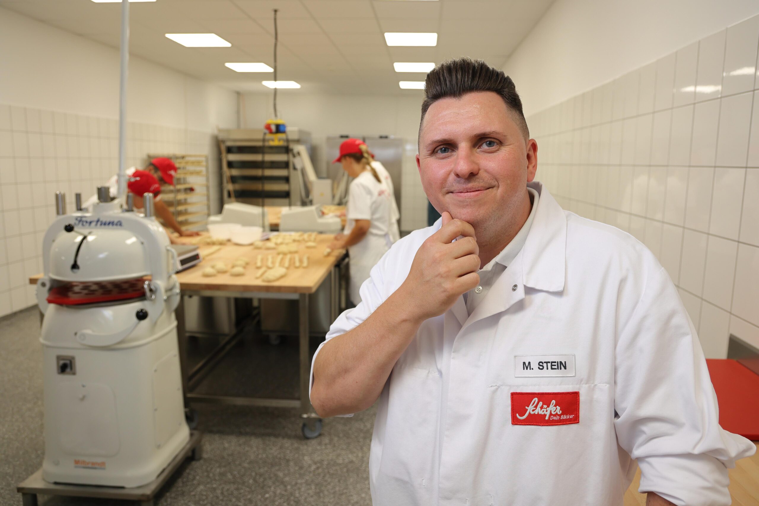 Michael Stein – Geschäftsführer Betrieb bei Schäfer Dein Bäcker Ein Herr ist in der Backstube von Schäfer Dein Bäcker, in Arbeitskleidung mit Namensschild mit der Beschriftung M. Stein, während im Hintergrund Mitarbeitende Teig vorbereiten.