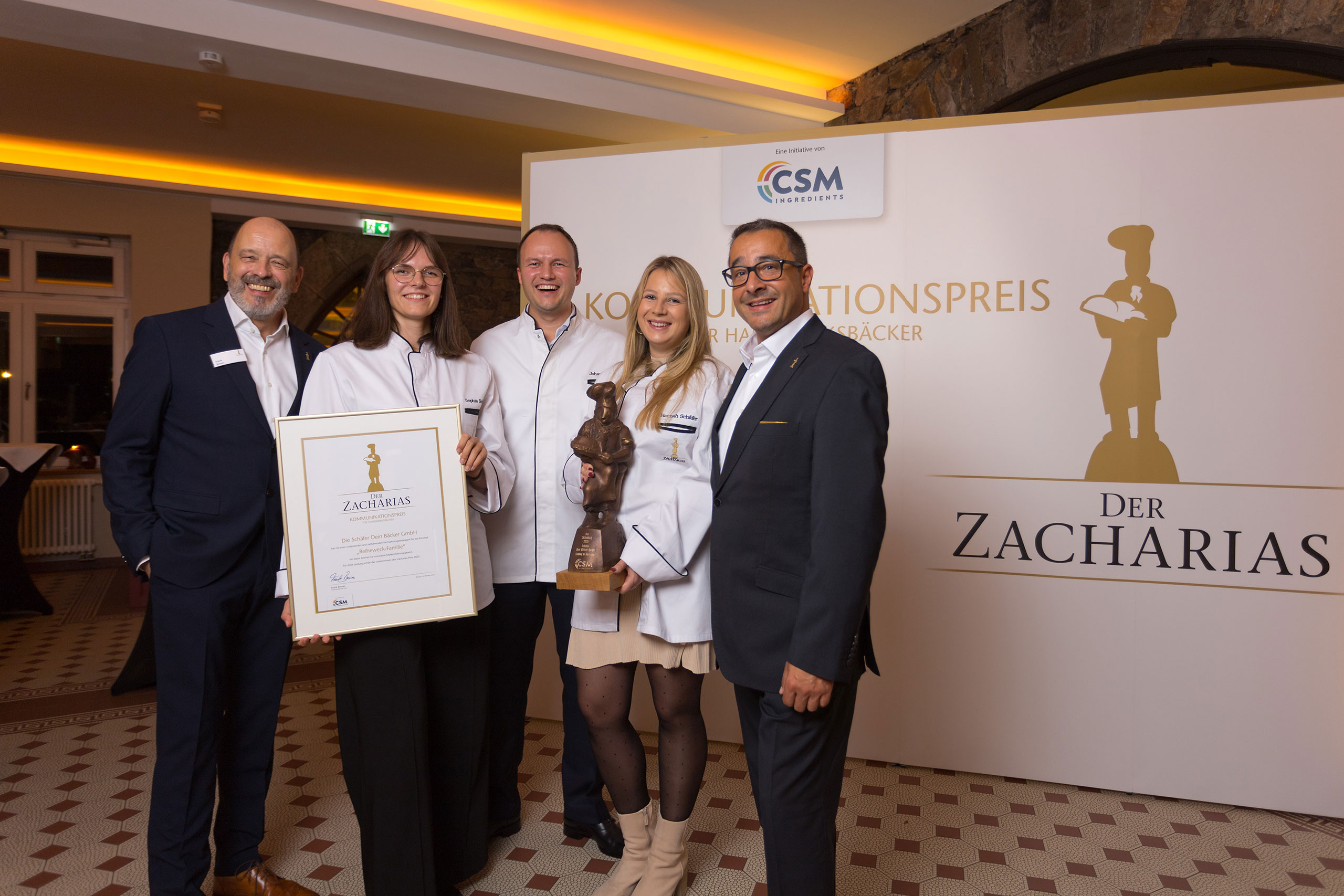 Zacharias 2025 — Auszeichnung Fünf Personen halten eine Urkunde und eine Zacharias-Trophäe vor einer Fotowand.